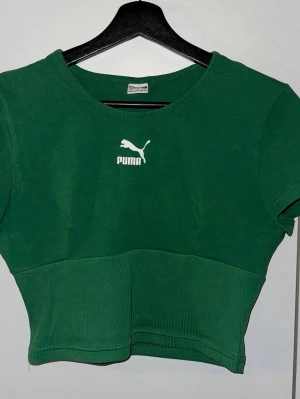 Grön croppad topp från Puma - Snygg grön croppad topp från Puma med vit logga framtill. Toppen har rund halsringning och korta ärmar. Nedre delen är ribbad för extra stilpoäng. Perfekt för dig som gillar sportig vibe och streetwear.