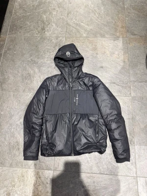 Svart pufferjacka med huva från Sail Racing - Säljer en svart pufferjacka från Sail Racing med huva och dragkedja framtill. Jackan har en cool paneldesign över bröstet och ryggen, samt en bröstficka med logotypdetalj. Perfekt för dig som gillar streetwear och vill hålla dig varm.