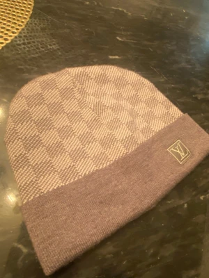 Rutig beige Louis Vuitton mössa - Snygg beige mössa från Louis Vuitton med rutigt mönster och uppvikt kant. LV-logga i hörnet ger en lyxig touch. Stickad i mjukt material som håller dig varm och stilren under kalla dagar.