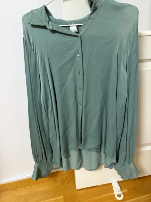 Grön skjorta med knappar, storlek M - Snygg grön skjorta i mjukt material med klassisk krage och knappar framtill. Skjortan har långa ärmar med breda manschetter och en loose passform som ger en chill vibe. Perfekt att styla med jeans eller kjol för en cool look.