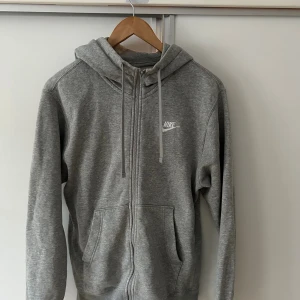 Grå hoodie från Nike med dragkedja - Grå hoodie från Nike med vit logga på bröstet, hel dragkedja och snörning i huvan. Tröjan är i storlek small men jag skulle säga att den är i M då den passar mig bra!