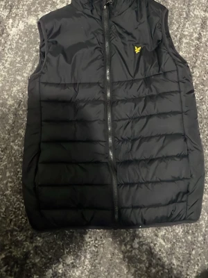 Svart dunväst från Lyle & Scott - Säljer en svart dunväst från Lyle & Scott med gul broderad logga på bröstet. Västen har quiltad design, rak passform och dragkedja framtill. Perfekt att slänga över en hoodie när det är kyligt ute. Materialet är syntet och dun.