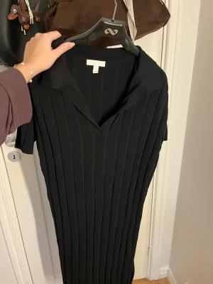 Svart ribbad klänning från H&M - Svart ribbad klänning med krage och korta ärmar från H&M. Klänningen har en v-ringad hals och är gjord i ett stickat material som ger en snygg struktur. Passformen är normal och längden går till knäna. Perfekt för dig som gillar stilrena och enkla plagg.