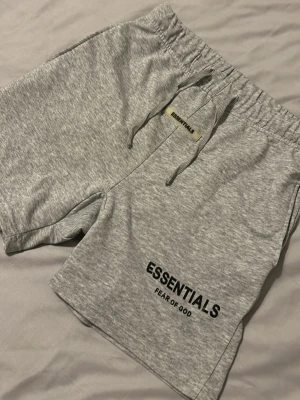 Grå Essentials shorts  - Säljer ett par grå mjuka shorts från Fear of God Essentials med svart logotyptryck på benet. De har elastisk midja med snörning och är tillverkade i bomullsmaterial. Perfekta för chill dagar eller träning.