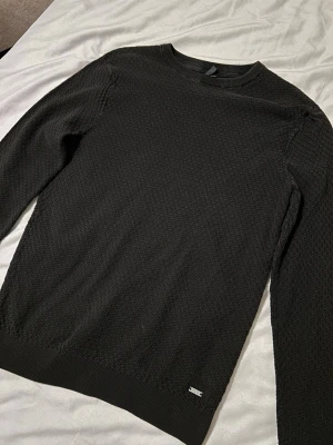 Svart stickad tröja med struktur - Snygg svart stickad tröja från Jack & Jones med diskret strukturerat mönster. Rund hals och långa ärmar, perfekt för lager-på-lager. Tröjan har en liten metallbricka vid nederkanten som detalj. Passar dig som gillar stilrena och enkla plagg.