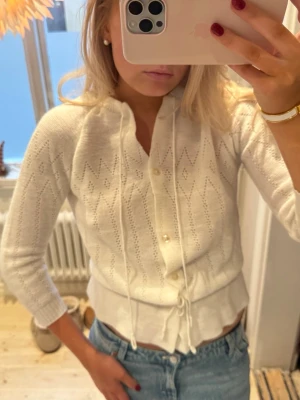 Vit stickad kofta med snörning - Superfin vit stickad kofta med snyggt hålmönster och knappar framtill. Koftan har snörning i midjan och vid halsen, samt trekvartsärm. Perfekt att styla med jeans för en chill look. Mjuk och lätt i materialet.