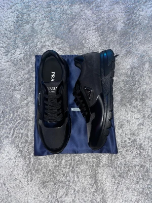 Svarta sneakers från Prada - Säljer ett par svarta Prada sneakers med chunky sula och snygga detaljer i skinn och textil. Skorna har snörning och den klassiska Prada-loggan på sidan. Perfekta för dig som vill ha en stilren och lyxig look med streetkänsla.!!!Kommer med kvitto!!!