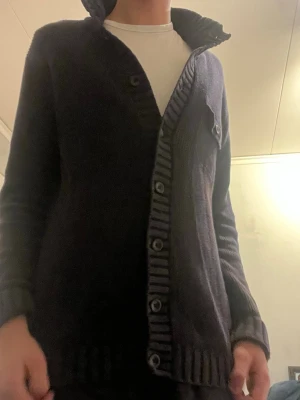 Mörkblå stickad kofta med knappar - Säljer en mörkblå stickad kofta med ribbade detaljer längs knappslån och nederkanten. Koftan har långa ärmar och en bröstficka med knapp. Perfekt för dig som gillar en chill och klassisk stil.