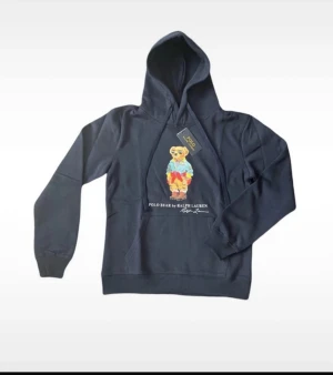 Polo Ralph Lauren Hoodie  - Helt ny! Säljer den här hoodien från Polo Ralph Lauren. Den har ett stort tryck mitt på bröstet med text under. Axelbredd 49cm, längd 63cm. JAG SKICKAR EJ FLER BILDER NÄR JAG BÄR JEANSEN! för storleksguide hänvisar jag till måtten och för bättre inblick i passformen rekommenderar jag att googla på modellnamnet. kolla måtten noggrant :) köpt på plick. 