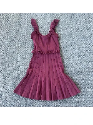 Vinröd plisserad klänning från Zara - Supersöt vinröd klänning från Zara med plisserad kjol och volangdetaljer på axelbanden och midjan. Klänningen är ärmlös och har en mjuk, stickad känsla i materialet. Perfekt för dig som gillar romantiska detaljer och vill sticka ut med färg.