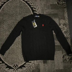 Svart stickad tröja Polo Ralph Lauren - Svart stickad tröja från Polo Ralph Lauren med klassisk kabelstickad design och röd broderad logga på bröstet. Rund halsringning och långa ärmar. Perfekt för dig som gillar stilrena och tidlösa plagg med en touch av lyx.