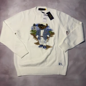 Vit stickad tröja Polo Ralph Lauren - Vit stickad tröja från Polo Ralph Lauren med ikonisk björn broderad på framsidan. Tröjan har rund hals, ribbade muddar och RL-broderi vid nederkanten. Materialet är bomull med detaljer i ull. Perfekt för dig som gillar klassisk och cool stil.