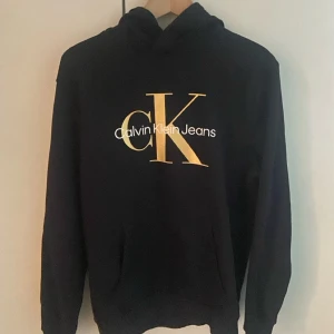 Svart hoodie från Calvin Klein Jeans - Svart hoodie från Calvin Klein Jeans med stor gul CK-logga och vit text på bröstet. Klassisk huva och magficka, perfekt för en chill och snygg look. Mjuk bomullskänsla och långärmad modell.