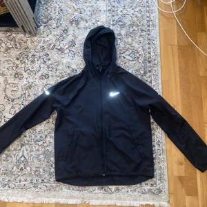 Svart Nike Miller vindjacka med huva - Svart vindjacka från Nike Running med reflekterande logga på bröstet och axeln. Jackan har huva, dragkedja framtill och två sidofickor. Tillverkad i lätt polyester, perfekt för löpning och utomhusaktiviteter. Stilren och sportig design som funkar året runt.