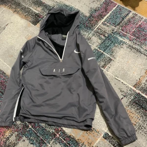Grå Nike Air vindjacka med huva - Snygg grå Nike Air vindjacka med huva och vita detaljer. Jackan har halv dragkedja framtill, stor ficka med AIR-tryck och meshfoder. Reflekterande tryck på ärm och rygg, samt elastiska muddar. Perfekt för dig som gillar sportig streetstyle.