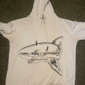 Vit hoodie med hajtryck från Palm Angels - Säljer en vit hoodie från Palm Angels med stor hajillustration på både fram- och baksidan. På ryggen finns även Palm Angels-loggan i gotisk stil. Hoodien har snörning vid huvan och är tillverkad i mjuk bomull. Perfekt för dig som gillar streetwear med unik design.