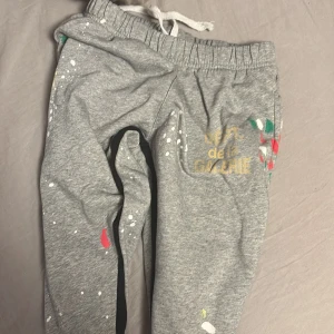 Grå mjukisbyxor från Gallery Dept. - Grå mjukisbyxor från Gallery Dept. med coolt färgstänksmönster och tryck i gult på ena benet. Byxorna har elastisk midja med snörning och är gjorda i mjukt bomullsmaterial. Perfekta för en chill och kreativ vibe.