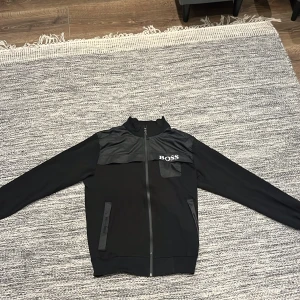 Svart kofta från Hugo boss, storlek M - Svart kofta från Hugo boss med stilren design och logga på bröstet. Koftan har hel dragkedja framtill, två fickor med detaljer i blankt material och hög krage. Materialmix med fleece och syntetpaneler upptill ger en modern look.