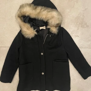 Svart parkajacka med fuskpäls från Zara  - Svart parkajacka från Zara med huva och fluffig fuskpäls runt kanten. Jackan har två stora fickor framtill, knappar och dragkedja. Klassisk stil. 