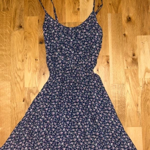 Blommig blå kortklänning Hollister XS - Supersöt blå klänning från Hollister i storlek XS med små rosa och vita blommor över hela tyget. Klänningen har smala axelband, snörning i ryggen och en lätt utställd kjol. Perfekt för sommarens alla tillfällen!