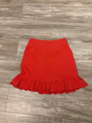 Röd volangkjol med snörning - Snygg röd kortkjol från VERO MODA med volangkant längst ner och snörning i midjan. Kjolen är gjord i mjuk bomull och har en enkel, feminin design som ger en cool vibe. Perfekt för dig som gillar färg och vill sticka ut lite extra.