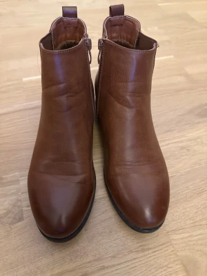 Bruna chelsea boots i skinn, strl 38 - Snygga bruna chelsea boots i skinn med rund tå och platt sula. Bootsens klassiska design har resår på sidorna och dragkedja på insidan för enkel på- och avtagning. Perfekt till höstens och vinterns outfits, passar dig som gillar stilrena och tidlösa skor.