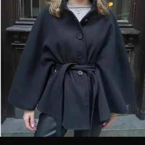 Svart poncho med bälte och knappar - Snygg svart poncho med breda ärmar och knytbälte i midjan. Perfekt för kyliga dagar. Enkel och clean design som funkar till många stilar.