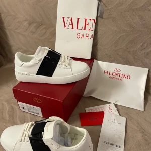 Valentino Garavani sneakers - Säljer ett par helt nya och oanvända Valentino Garavani sneakers i vitt skinn med en bred svart rem över vristen och ikoniska nitar på hälen. Klassisk rund tå, platt sula och snygga detaljer. Kommer med originalkartong, dustbag och tags. Säljs pga fel storlek