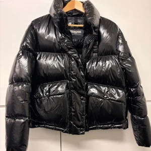 Svart pufferjacka från Blauer - Snygg svart pufferjacka från Blauer med glansig finish och hög krage. Jackan har stora fickor framtill, Blauer-logga på ärmen och dragkedja med tryckknappar. Perfekt för kalla dagar när du vill hålla stilen och värmen.
