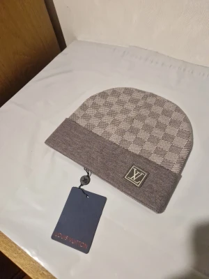 Louis Vuitton rutig grå mössa - Snygg grå mössa från Louis Vuitton med klassiskt rutigt mönster och LV-logga framtill. Mössan har uppvikt kant och är tillverkad i mjukt stickat material som känns lyxigt och bekvämt. Perfekt för dig som vill ha en stilren accessoar med designer-vibe.
