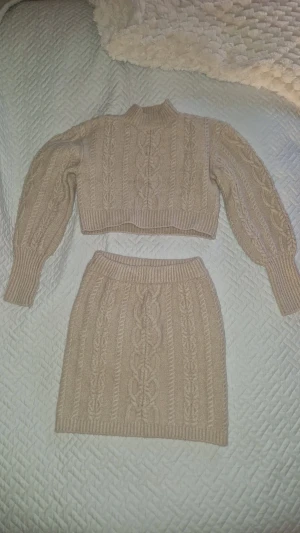 Klädset - Superfin beige stickad kortkjol med bred resår i midjan och en matchande fin tröja, båda med snyggt flätmönster. Det är i mjukt material och har en klassisk, ribbad kant nertill. Perfekt för en mysig look. Sparsamt använt- inga synliga defekter utöver en liten loop som dragits ut lite på insidan- se bild. Inget som syns på utsidan. Två olika storlekar men passat mej som storlek S