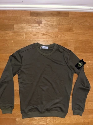 Olivgrön Stone Island sweatshirt L - Snygg olivgrön sweatshirt från Stone Island i storlek L. Tröjan har rund hals, ribbade muddar och den klassiska Stone Island-loggan på vänster ärm. Materialet är mjuk bomull och passformen är normal. Perfekt för dig som gillar streetwear och vill ha en clean look.