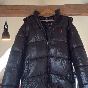 Svart pufferjacka från Polo Ralph Lauren | Vinter jacka - Svart pufferjacka från  Ralph Lauren med glansig finish och  röd logga på bröstet. Köpte jackan förra vintern och användes bara när det var som kallast, fortfarande i så gått som nyskick. Syns absolut inte att den är använd på något sätt. Mvh Anton.