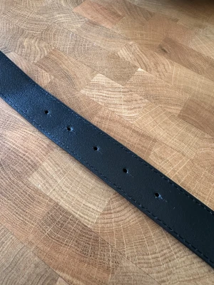 Svart skärp från J.Lindeberg i skinn - Snyggt svart bälte från J.Lindeberg i äkta skinn, tillverkat i Italien. Bältet har en unik, geometrisk silverfärgad spänne som ger en modern vibe. Perfekt för dig som vill ha en stilren accessoar med lite edge.