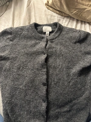 Mörkgrå kofta från H&M XS - Mysig mörkgrå kofta från H&M i storlek XS. Koftan har rund halsringning, långa ärmar och knäppning med svarta knappar framtill. Materialet är stickat och känns mjukt mot huden, perfekt för kyliga dagar.