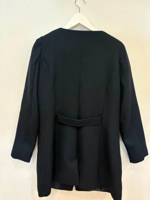 Svart rock från Jyothi med dragkedja - Stilren svart rock från Jyothi med rund halsringning och silverfärgade dragkedjor framtill samt vid fickorna. Jackan har lång ärm och ett bälte baktill för extra detalj. Materialet är strukturerat och ger en snygg look.