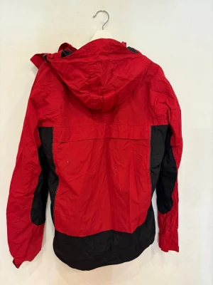 Röd och svart vindjacka från Clique - Snygg röd och svart vindjacka från Clique med huva och flera praktiska fickor. Jackan har dragkedja och kardborreknäppning framtill, samt justerbar huva med snörning. Perfekt för blåsiga dagar och outdoor-aktiviteter.