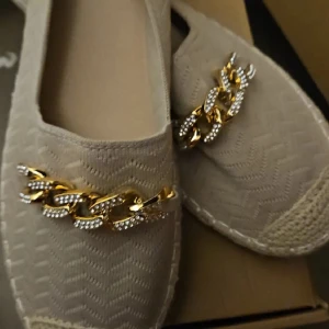 Beige ballerinaskor med guldkedja - Snygga beige ballerinaskor med rund tå och platt sula. Skorna har ett stickat mönster och är dekorerade med en glittrig guldfärgad kedja framtill. Perfekta för dig som gillar detaljer och vill ha en unik touch på din outfit.