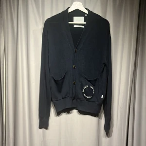 Mörkblå kofta från Les Deux - Snygg mörkblå kofta från Les Deux med broderad logga på fickan. Koftan har v-ringad hals, tre knappar framtill och två fickor. Materialet är stickat och ger en chill och stilren vibe. Perfekt för lager-på-lager och passar till jeans eller chinos. Nypris 1000kr