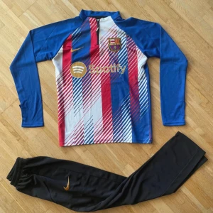 Barcelona långärmad fotbollströja Nike - Barcelona långärmad fotbollströja från Nike med klubbmärke och Spotify-logga på bröstet. Tröjan har ett randigt mönster i blått, rött och vitt med guldfärgade detaljer. Materialet är lätt och andas, perfekt för träning eller match.