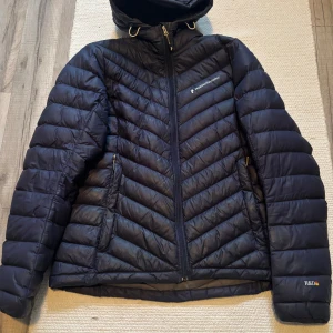 Mörkblå Peak Performance dunjacka L - Snygg mörkblå dunjacka från Peak Performance i storlek L. Jackan har huva med dragsko, två sidofickor med dragkedja och coolt quiltat mönster. jackan har en liten lagad reva på armen som knappt syns och dragkedjan kan vara lite kärv om man drar upp den oförsiktigt Perfekt för kalla dagar och har en sportig vibe. Logga på bröstet och ärmen.