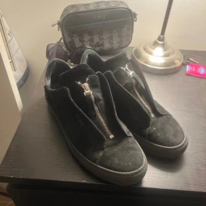 Arigato skor zip - Svarta sneakers i mocka från ARIGATO med cool dragkedja framtill istället för klassisk snörning. Rund tå och platt sula ger en clean och modern vibe. Perfekt för dig som gillar minimalistisk stil med en twist. Finns lite tecken på användning pgd moped växelspaken.