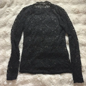 Svart spetsblus med lång ärm från Gina genomskinlig  - Säljer en svart blus i genombruten spets med lång ärm och rund hals. Blusen har ett detaljerat blommigt spetsmönster och är transparent, vilket ger en snygg och edgy vibe. Perfekt att styla med jeans eller kjol för en cool look. Den går upp lite mot halsen och är genomskinlig 