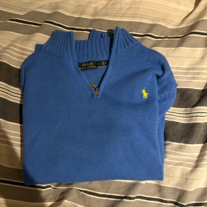 Blå stickad tröja Polo Ralph Lauren - Blå stickad tröja från Polo Ralph Lauren med gul broderad logga på bröstet. Tröjan har ribbad krage och muddar, samt en half zip-dragkedja vid halsen. Perfekt för dig som gillar klassisk stil med en sportig touch.