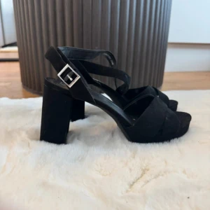 Svarta sandaler med klack och spänne - Snygga svarta sandaler med bred klack och öppen tå. Remmar i mocka-look som korsas över foten och ett silverfärgat spänne vid ankeln. Perfekta för dig som vill ha både höjd och stabilitet. Plattformsula för extra komfort.