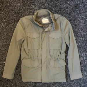 Grön field jacket - Snygg grön parkajacka från East West med fyra stora fickor framtill och hög krage med tryckknappar. Jackan har dragkedja och meshfoder på insidan. Perfekt för dig som gillar funktionell och stilren design.