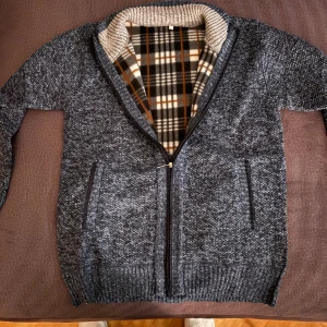 Mörkgrå/blå kofta med dragkedja - Mysig mörkgrå/blå kofta med hel dragkedja och ribbad krage. Insidan har ett rutigt foder i svart, vitt och brunt som ger extra värme och stil. Två fickor framtill och långärmad modell, perfekt för kyliga dagar.fickorna är bara disign! Helt ny 