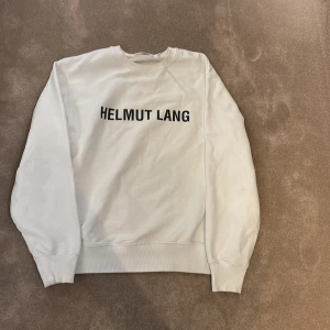 Vit sweatshirt från Helmut Lang - Vit sweatshirt från Helmut Lang med svart tryck på bröstet. Tröjan har rund hals, ribbade muddar och är långärmad. Materialet är mjuk bomull som känns skönt mot huden. Perfekt för dig som gillar stilrena och minimalistiska plagg.