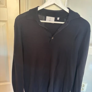 Svart pikétröja från Bläck - Klassisk svart pikétröja från Bläck med lång ärm och krage. Tröjan har två knappar framtill och är gjord i mjukt bomullsmaterial som känns skönt mot huden. Perfekt för dig som gillar stilrena och enkla plagg med snygg passform.