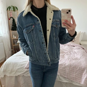 Levi's jeansjacka med teddyfoder - Säljer en klassisk blå jeansjacka från Levi's med mjukt vitt teddyfoder och krage som är perfekt för lite kallare väder nu. Jackan har två bröstfickor med knappar, sidofickor och knappstängning framtill. Perfekt för dig som gillar retrovibe och vill ha något som är både bekvämt och snyggt. Den är nästan oanvänd då den bara använts ett fåtal gånger och sen tvättats innan den hängdes in i garderoben. Den är varm och skön men lite större i storleken så de ger en lite oversized look. Nypris- 1600:-